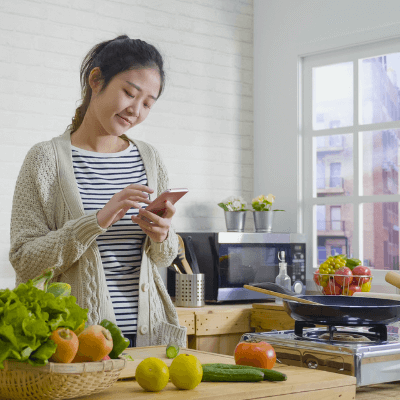 Young Asian woman cooking vegetables in wooden kitchen while looking at recipe on smartphone. 
